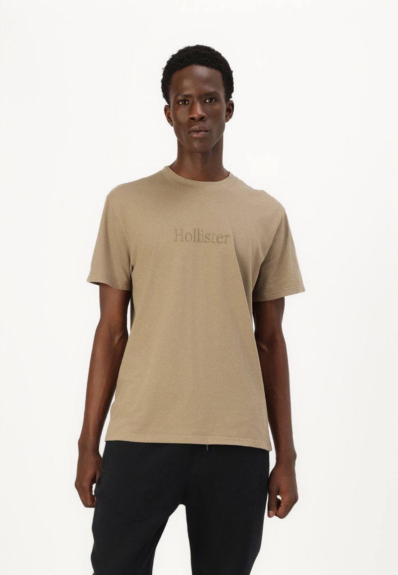 Bežinė medvilninė marškinėliai trumpomis rankovėmis su apvaliu iškirpimu, su subtiliu siuvinėtu "Hollister" logotipu ant krūtinės.