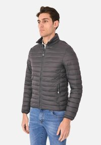 Giubbotto di piumino grigio con cuciture orizzontali, chiusura con zip, colletto alto e tasche laterali. Indossato sopra una camicia con colletto e jeans.