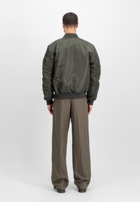 Olive green bomber jacket with ribbed collar and cuffs, featuring padded shoulders and elastic waistband, paired with gray pleated trousers.