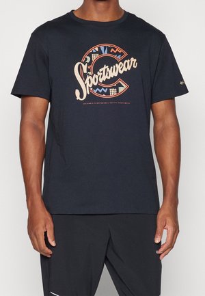 Mies, jolla on musta t-paita, jossa on värikkäät "Sportswear"-teksti ja kuviot ison C-kirjaimen sisällä rinnassa, sekä mustat housut.