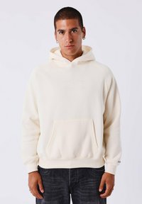 Jonge man met kort haar, gekleed in een crèmekleurige hoodie met een voorzak en een zwarte jeans, staande tegen een effen witte achtergrond.