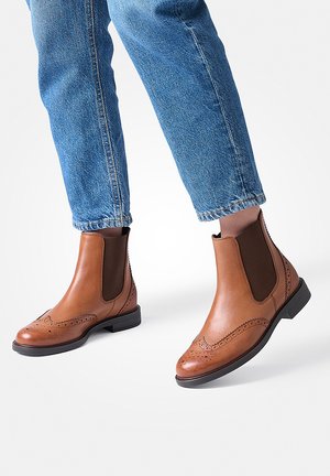 Person iført lyseblå denim jeans og brune læder Chelsea-støvler med brogue detaljer, stående på en hvid overflade.