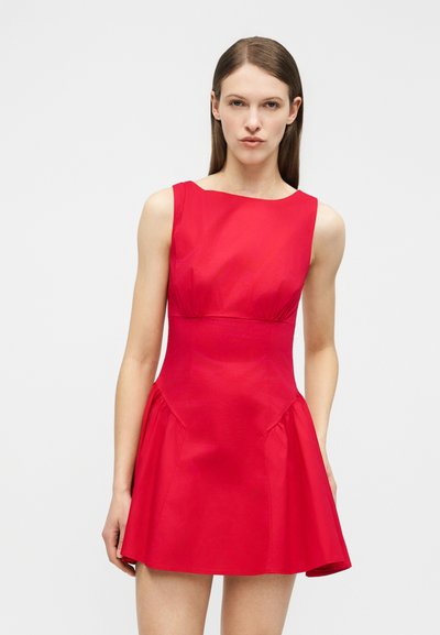 Γυναίκα με ίσια καστανά μαλλιά που φοράει αμάνικο κόκκινο φόρεμα με φούστα που ανοίγει, στέκεται μπροστά σε απλό ανοιχτό γκρι φόντο.