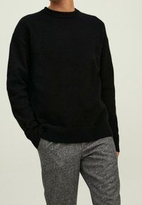 Pull noir en tricot avec un col rond, des manches longues et des poignets côtelés ; associé à un pantalon gris texturé.