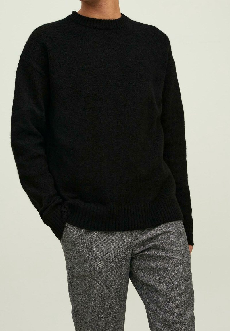 Pull noir en tricot avec un col rond, des manches longues et des poignets côtelés ; associé à un pantalon gris texturé.