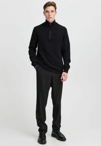 Pulover negru tricotat cu guler înalt și fermoar pe jumătate, purtat cu pantaloni negri de birou și pantofi negri lustruiți. Textură netedă.