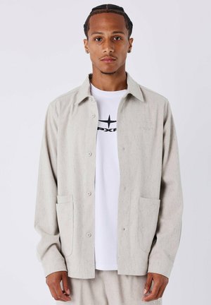 Joven con el cabello trenzado que lleva una chaqueta beige de botones sobre una camiseta gráfica blanca, de pie frente a un fondo blanco liso.