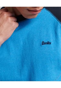 Sininen puuvillapaita, jossa on musta kaula-aukko, jossa on pieni brodeerattu "Superdry" -logo rintamuksessa. Sileä pinta, rento muotoilu.