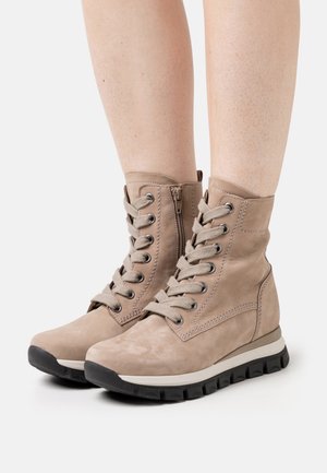 Beige veterlaarzen tot aan de enkel met zijritsen en zwarte rubberen zolen, gedragen op blote benen tegen een lichte achtergrond.