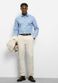 Hellblaues Hemd mit Knöpfen und einem dezenten Muster, kombiniert mit cremefarbenen Hosen, einem beigen Blazer über einem Arm drapiert und dunklen Anzugschuhen.