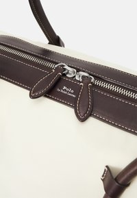 Beżowa torba z płótna z ciemnobrązowymi skórzanymi akcentami, zamykanie na zamek błyskawiczny u góry oraz etykieta z logo. Posiada podwójne skórzane uchwyty i kontrastowe szwy.