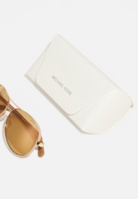 Michael Kors solbriller med brune linser og tynne gullrammer, akkompagnert av et strukturert hvitt etui med logo.