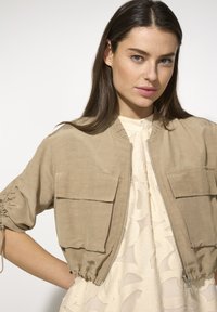 Beige gefütterte Bomberjacke mit kurzen Puffärmeln, zwei Fronttaschen und Reißverschluss, aus leichtem Stoff mit einer glatten Textur.