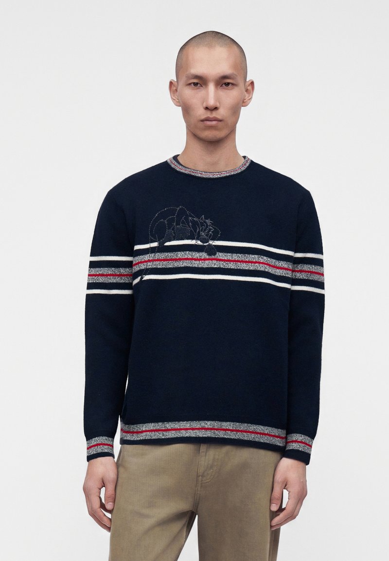Homme portant un pull bleu marine avec des rayures horizontales blanches, rouges et grises et un petit chat de dessin animé brodé sur la poitrine, associé à un pantalon beige.