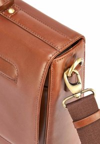 Brązowa skórzana torba o gładkim wykończeniu, z elementami ze złotego metalowego, odpinanym canvasowym paskiem oraz detalami w postaci przeszycia wzdłuż krawędzi.