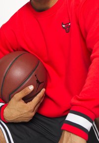Červená mikina s černým logem Chicago Bulls, nošená přes černé kraťasy s bílými a červenými pruhovanými akcenty. Drží texturovaný basketbal.