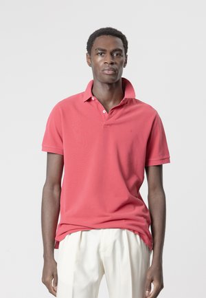 Hombre con camiseta tipo polo coral con cuello levantado y pantalones blancos, de pie frente a un fondo liso gris claro.