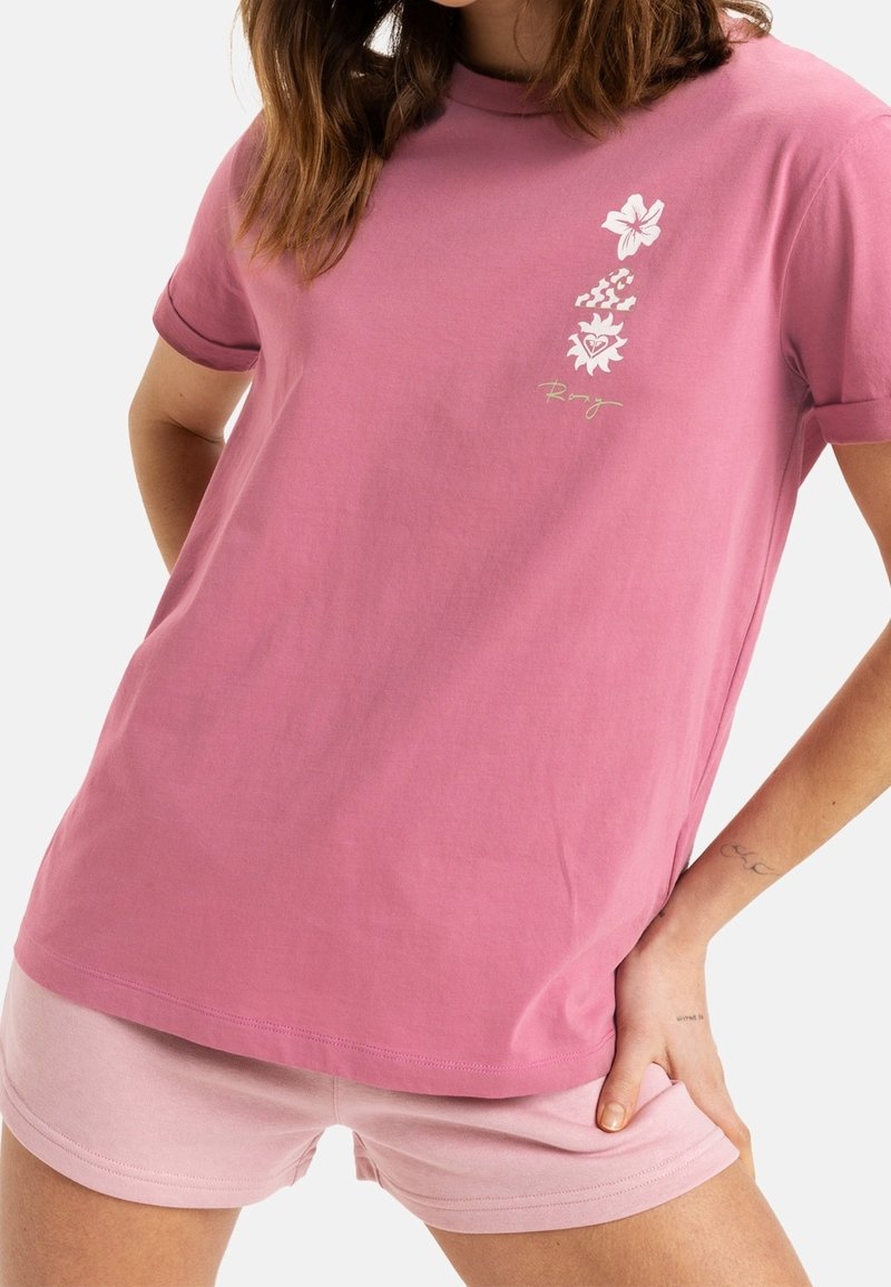 Žena v růžovém tričku se bílými květinovými a vlnovkovými logy, v kombinaci se světle růžovými šortkami, stojící s rukama v bok.