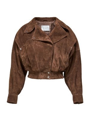 Vrouw met een kort bruin suède jasje, zwarte leren knie-lange rok, zwarte kniehoge laarzen, houdend een zwarte leren handtas vast.