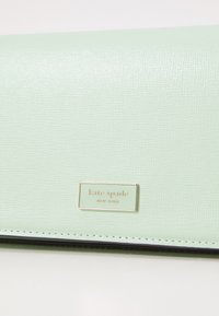 Mint green leather wallet with textured surface, featuring a gold logo plate and contrasting stitching along the edges.