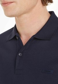 Mörkblå pikétröja av texturerad bomull, med klassisk krage, två knappar och en liten logglabel på bröstet.