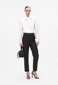 White long-sleeve shirt with a large bow collar, paired with dark textured trousers, black woven handbag, and strappy high-heeled sandals.