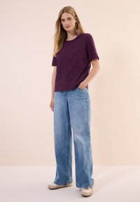 T-shirt di cotone viola con maniche corte, abbinato a jeans a gamba larga blu chiaro. Il modello sta in piedi con le mani in tasca, indossando scarpe beige.