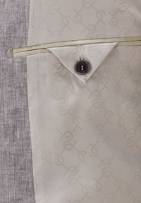 Hellbeige Stoff mit einem dezenten Logo-Muster, kombiniert mit strukturiertem grauem Material. Verfügt über eine knopfbeschlossene Tasche mit einem kontrastierenden Besatz.