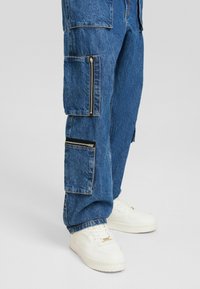 Pantalones cargo de mezclilla azul con múltiples bolsillos grandes y cierres de cremallera, combinados con zapatillas blancas. La tela tiene una textura desgastada.