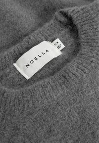 Weiches, graues gestricktes Kleidungsstück mit weißem Etikett "NOELLA" und Größenschild "S/M" sowie dem Hinweis "Made in Italy" im Kragenbereich.