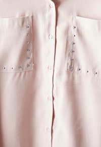 Light pink button-up shirt with two front pockets, adorned with silver studs along the pockets. Smooth fabric and classic collar design.