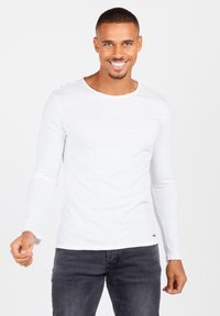 Maglietta bianca a maniche lunghe realizzata in tessuto leggero, con scollatura rotonda e dettagli di cucitura laterale, abbinata a jeans scuri.