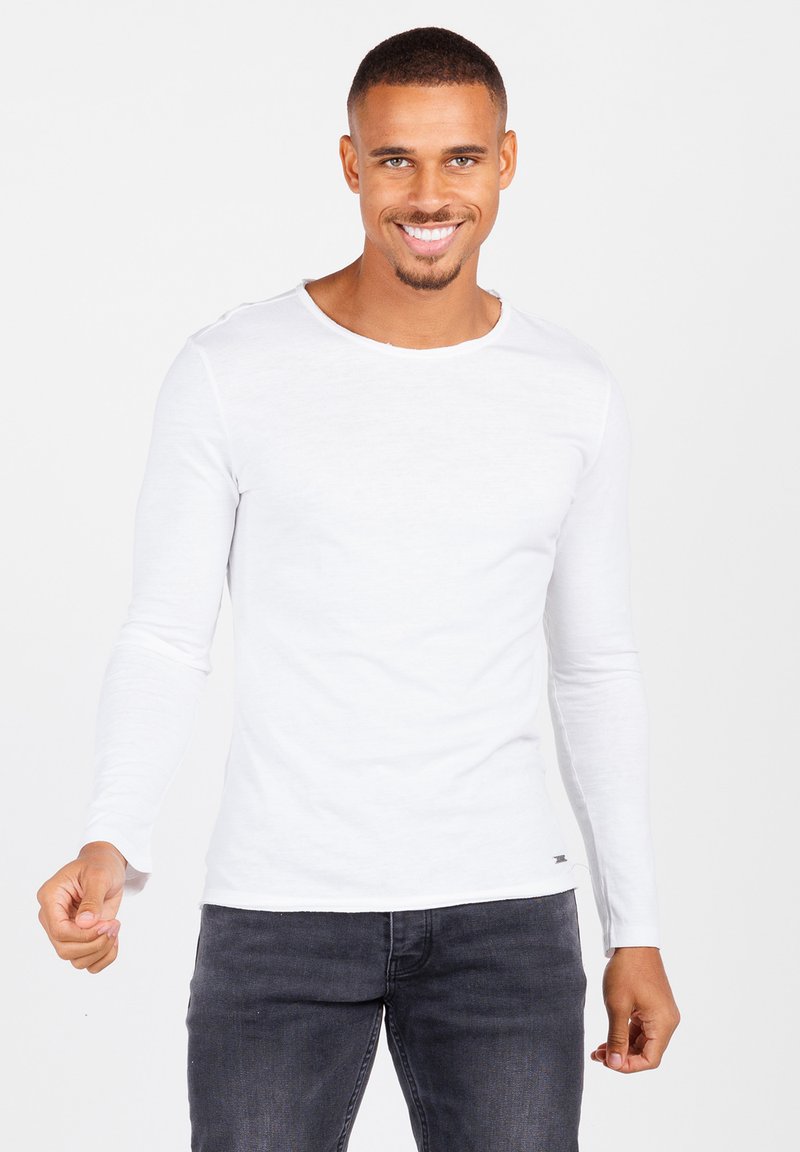 Maglietta bianca a maniche lunghe realizzata in tessuto leggero, con scollatura rotonda e dettagli di cucitura laterale, abbinata a jeans scuri.