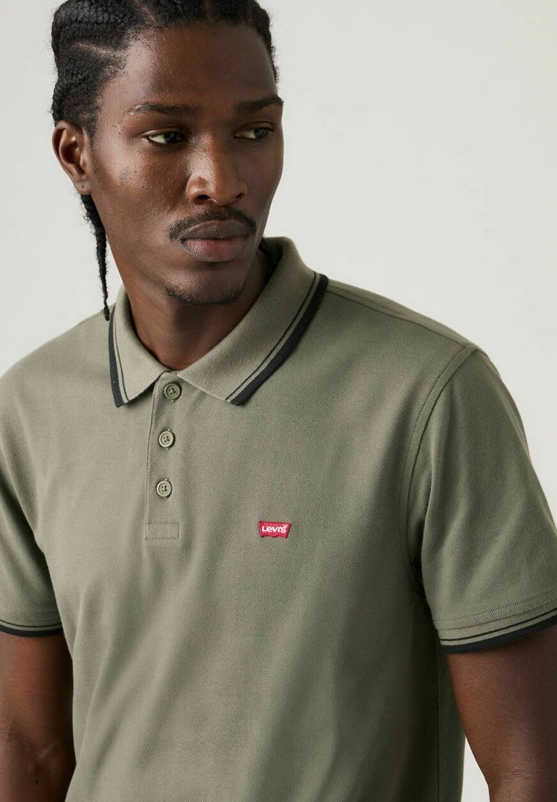 Polo verde oliva in cotone, con una patta a tre bottoni, rifiniture nere sul colletto e sulle maniche, e un logo Levi's rosso sul petto.
