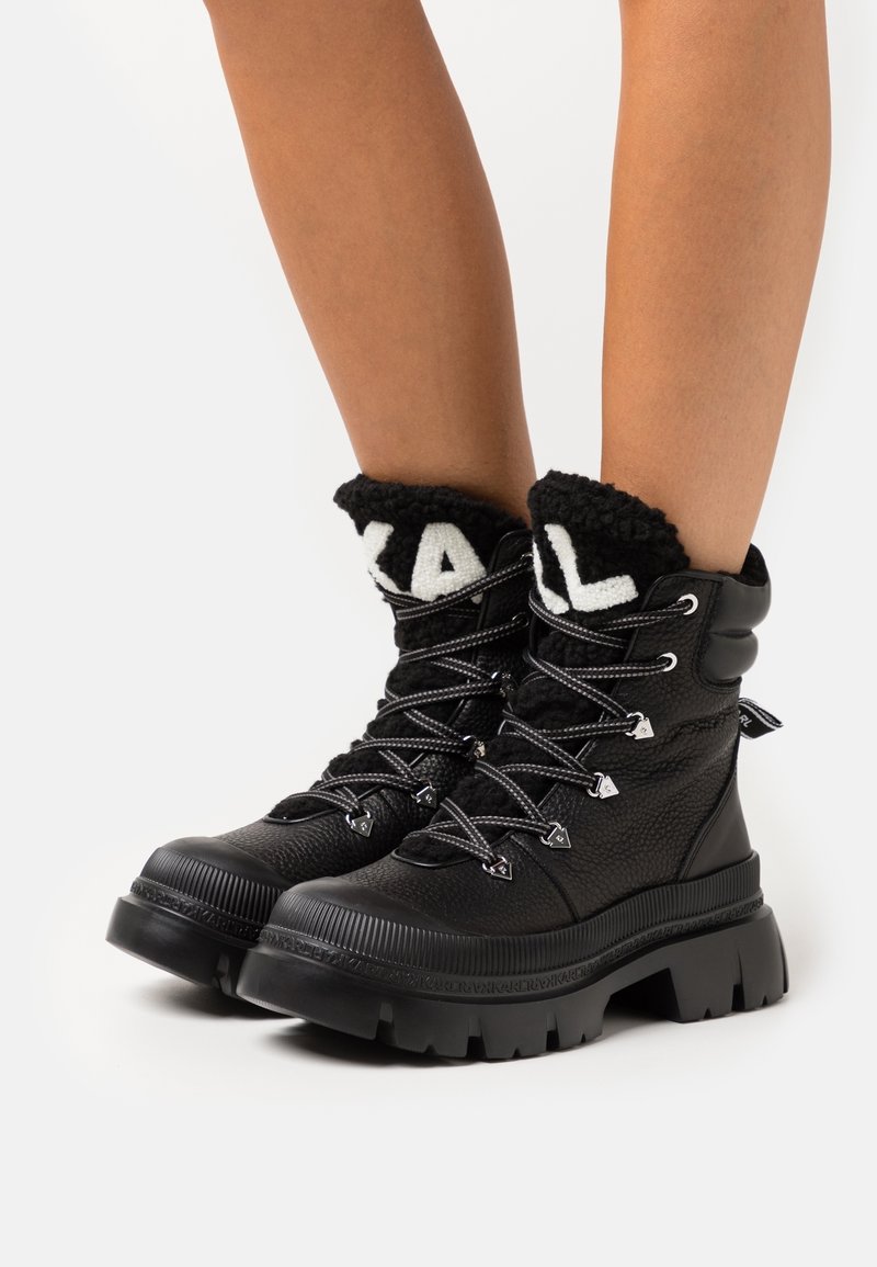 Botas de tobillo de cuero negro texturizado con suelas gruesas, parte delantera con cordones y puños esponjosos que presentan letras blancas en negrita.