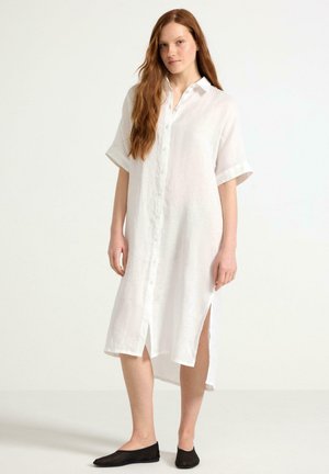 Abito camicia bianco in lino con maniche corte, fronte con bottoni, spacchi laterali e vestibilità comoda. Indossato con ballerine nere.