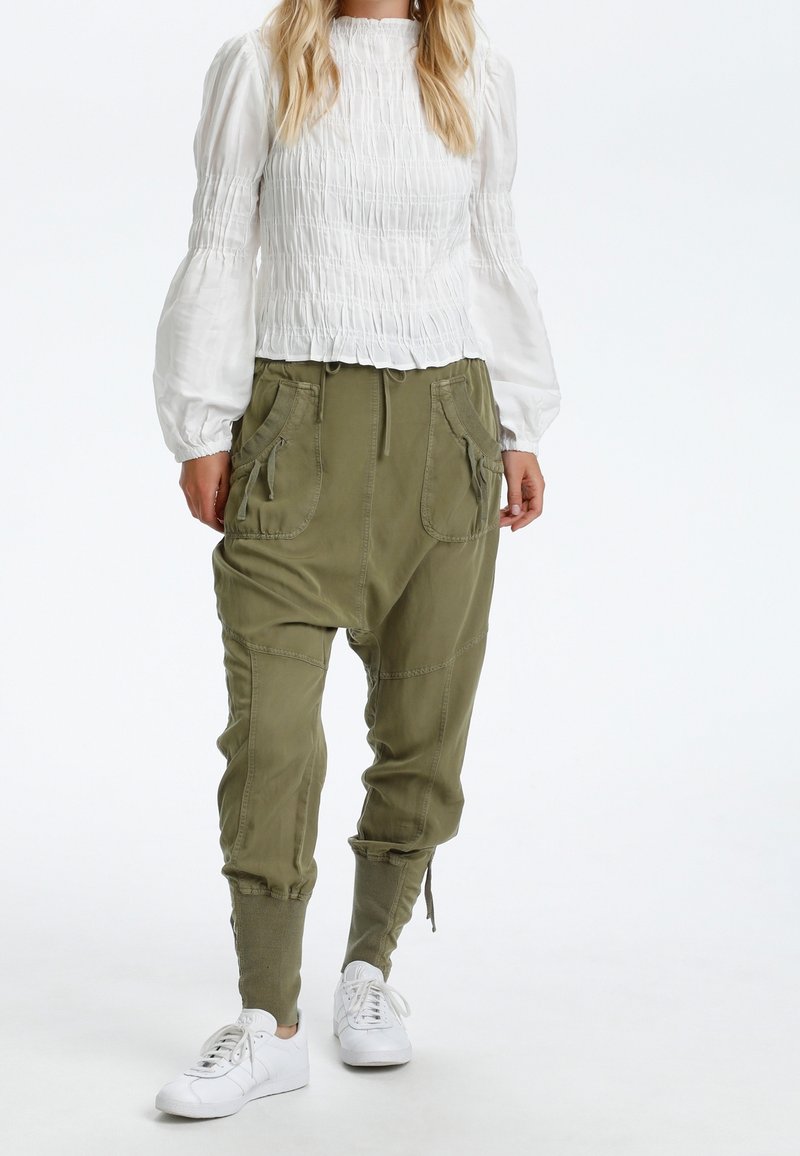 Haut blanc à manches longues avec smocks, associé à un pantalon ample vert olive avec poches latérales et poignets fuselés, porté avec des baskets blanches.