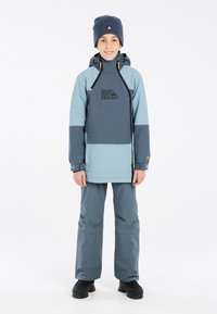 Veste imperméable bleue et grise avec une fermeture éclair à l'avant et une capuche, arborant un logo sur la poitrine, assortie à un pantalon et des chaussures noires.
