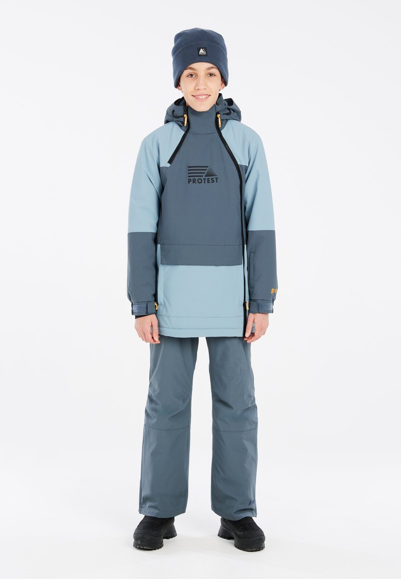 Veste imperméable bleue et grise avec une fermeture éclair à l'avant et une capuche, arborant un logo sur la poitrine, assortie à un pantalon et des chaussures noires.