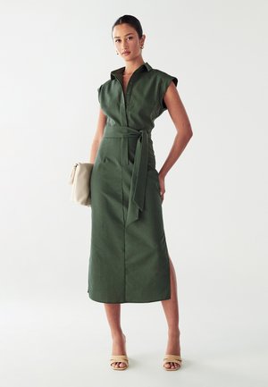 Vrouw in een mouwloze olijfgroene midi-jurk met een riem, die een beige clutch vasthoudt, draag beige open teen sandalen en gouden oorbellen, staande tegen een witte achtergrond.