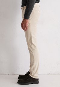Beige katoenen broek met een slim fit, voorzien van zijzakken en een subtiele textuur. Gepaard met donker suède schoenen, waardoor een gestroomlijnd profiel zichtbaar is.
