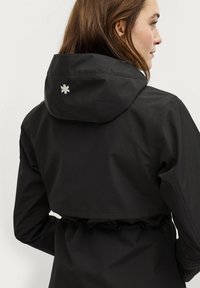 Giacca nera impermeabile con cappuccio, vita elastica e un dettaglio decorativo in ricamo bianco sulla schiena. Finitura in tessuto liscia.
