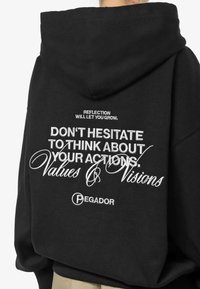 Svart huvtröja med en stor grafik på ryggen med vit text: "TVEKA INTE ATT TÄNKA PÅ DINA HANDLINGAR. Värderingar och Visioner."