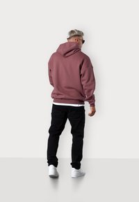 Rose-colored hoodie with a large hood, ribbed cuffs, and hem. Paired with black pants and white sneakers, emphasizing a casual look.