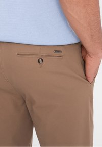 Mężczyzna w beżowych spodniach z guzikiem w tylnej kieszeni i jasnoniebieskiej koszuli, z ręką częściowo w kieszeni przedniej.