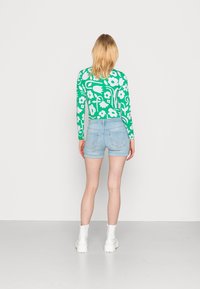 Top verde de manga longa com padrão floral branco, combinado com calções de ganga claros. Modelo usa botas brancas até ao tornozelo, posando contra um fundo simples.