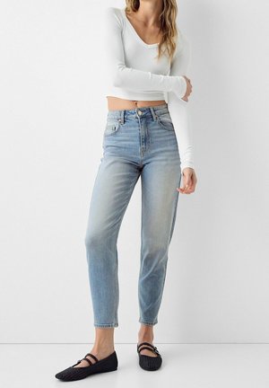 Kobieta w wysokich jasnoniebieskich dżinsach, białym długim topie odsłaniającym brzuch i czarnych plecionych balerinach, stojąca na tle jednolitego białego tła.