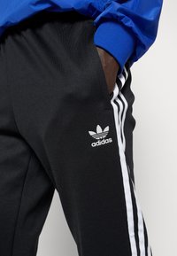 Černé sportovní kalhoty s bílými pruhy po stranách a nášivkou s logem Adidas. Hladká látka, elastický pas s kapsami. V horní části viditelná modrá bunda.