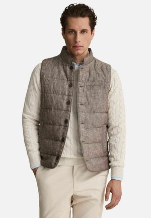 Man som bär en brun quiltad väst över en krämfärgad kabelstickad tröja och beige byxor, stående med ena handen i fickan.