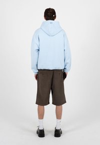 Homme de dos portant un sweat à capuche bleu clair, un short marron foncé jusqu'aux genoux, des chaussettes blanches, des chaussures noires et une casquette sombre, sur un fond uni.